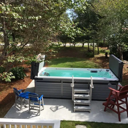Outdoor swim spa with clear water surrounded by chairs and trees on a sunny day.