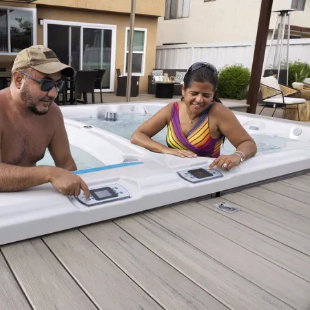 Man and woman adjusting controls while sitting in outdoor hot tub on wooden deck in backyard.