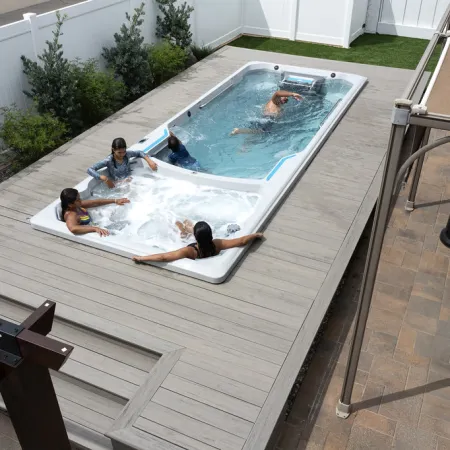 Four people enjoying a backyard swim spa with a swim area and jacuzzi section on a wooden deck under a canopy.