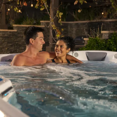 Couple smiling and enjoying a relaxing evening in a bubbling outdoor hot tub surrounded by garden lights.