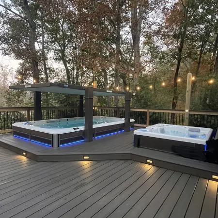 Modern backyard deck with two illuminated hot tubs surrounded by trees and ambient string lights at dusk.