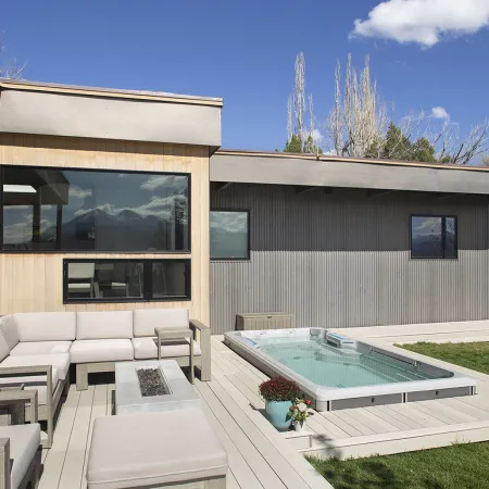Modern backyard with beige sectional sofa, built-in hot tub, fire pit, and gray wooden house exterior.