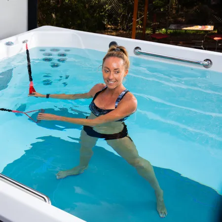 Woman exercising with resistance bands in a swim spa pool under bright lighting outdoors.