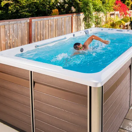 Man swimming in a modern outdoor swim spa with wooden paneling surrounded by garden and fence.