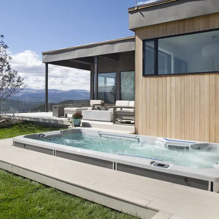 Modern backyard hot tub with bubbling water next to wooden house and patio overlooking mountain landscape.