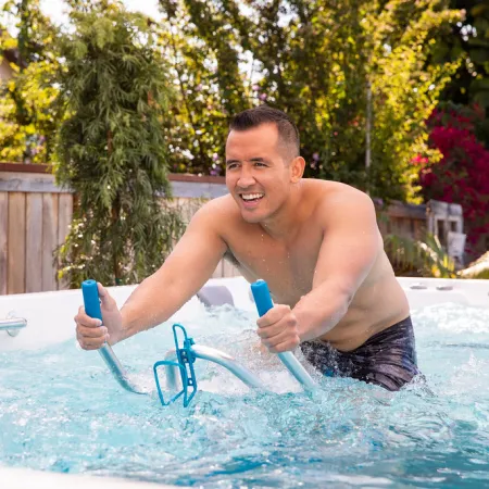 Man exercising on aquatic stationary bike in backyard hot tub with wooden fence and green trees.