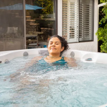 Woman relaxing with eyes closed in an outdoor hot tub surrounded by greenery and patio furniture.