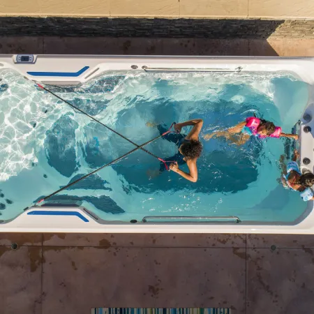 Overhead view of a person exercising with resistance bands in a swim spa while two children and an adult watch from the side.