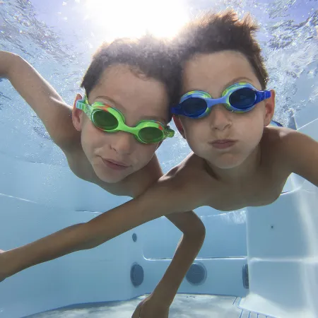 Two boys wearing swimming goggles underwater in a pool, smiling and reaching toward the camera