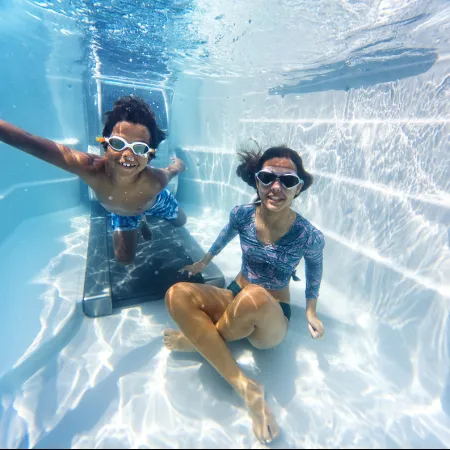 Two children wearing goggles underwater in a swimming pool playing near a treadmill submerged in water.