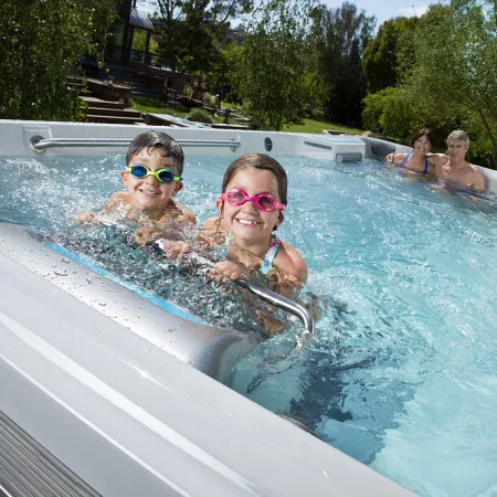 Two children with goggles and two adults relaxing in a backyard outdoor hot tub on a sunny day.