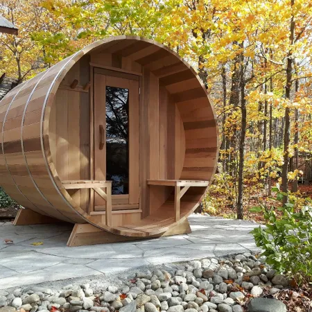 Round wooden barrel sauna with glass door on stone patio surrounded by autumn trees and fallen leaves.