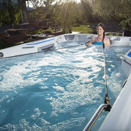 Woman exercising in a spacious outdoor swim spa with resistance handles and bubbles under sunlight.