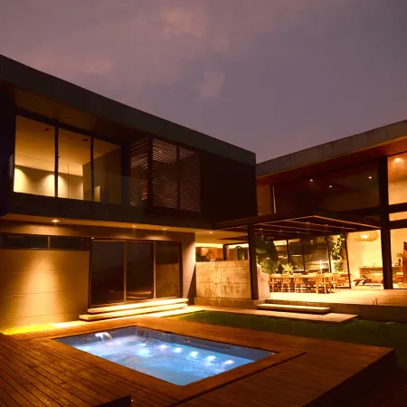 Modern two-story house with large glass walls, warm interior lighting, and illuminated outdoor pool at dusk.