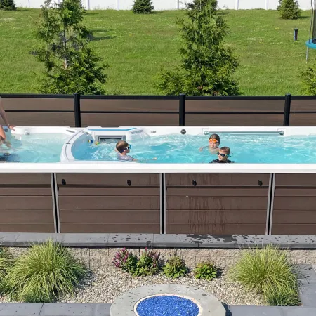 Family enjoying a modern above-ground swim spa in a sunny backyard with greenery and patio landscaping.