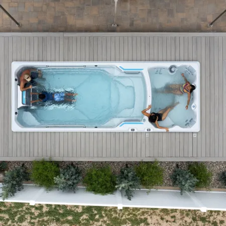Top view of an outdoor swim spa with three people relaxing and exercising in the water, surrounded by decking and garden.