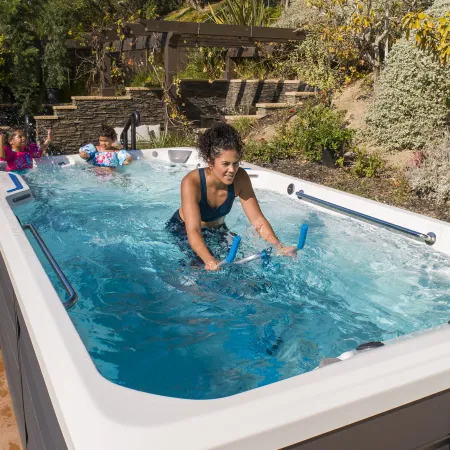 Woman exercising on water bike in backyard swim spa while two kids play in the water behind her