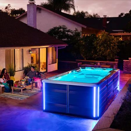 Evening backyard scene with a family relaxing next to a glowing blue-lit swim spa outside a cozy house.