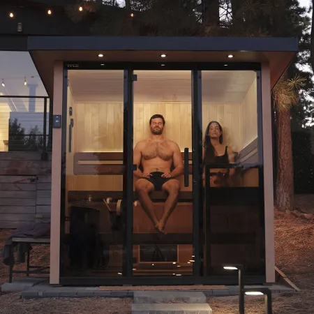 Man and woman relaxing inside a modern glass sauna cabin at dusk outdoors near trees.