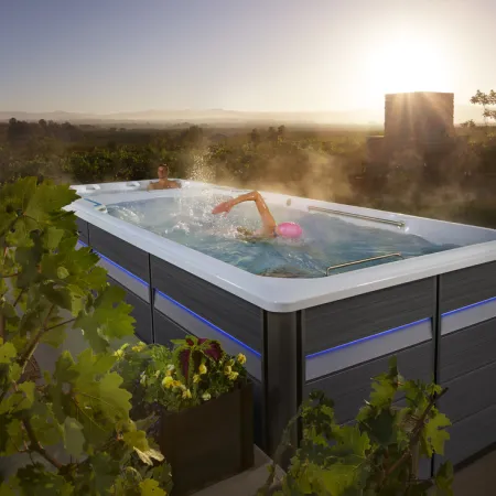 Outdoor swim spa with two people, one swimming and one relaxing, surrounded by plants at sunset.