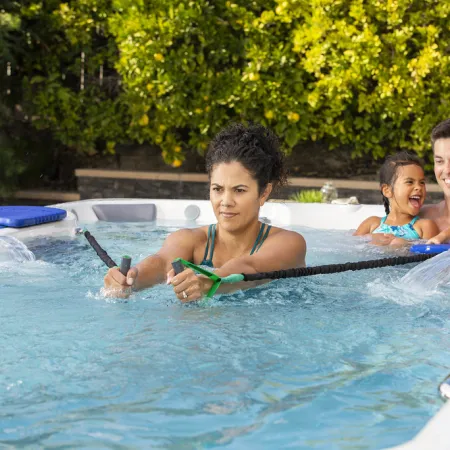 Woman exercises with resistance bands in a swim spa while a man and two children enjoy the water nearby.