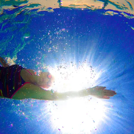 Underwater view of a swimmer reaching forward with sun shining brightly through blue water and bubbles around.