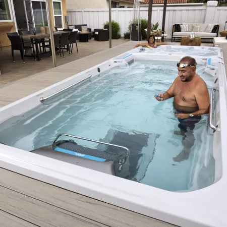 Man exercising in a swim spa pool with a woman relaxing in the background on a patio deck.