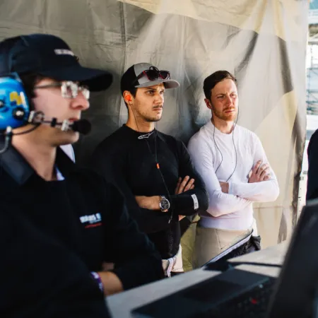 Four race engineers and team members monitoring data on screens during a motorsport event pit stop.