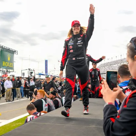 Racing driver in black and red suit and cap waves to crowd while walking on platform at race track event.