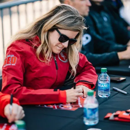 Person in red jacket and sunglasses signing autographs at an outdoor event table with water bottles.