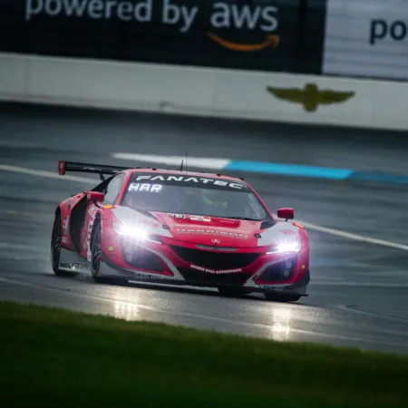 Red racing car with headlights on speeding on wet race track under overcast sky