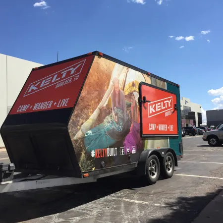 Kelty branded camper trailer with outdoor adventure graphics parked in a sunny parking lot under blue sky.