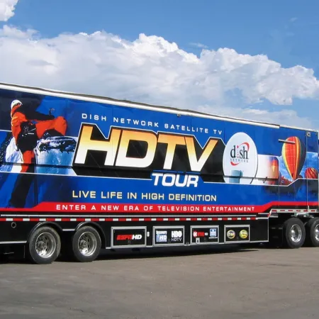 Large white semi-truck with a colorful trailer advertising DISH Network HDTV Tour with sky and clouds background.