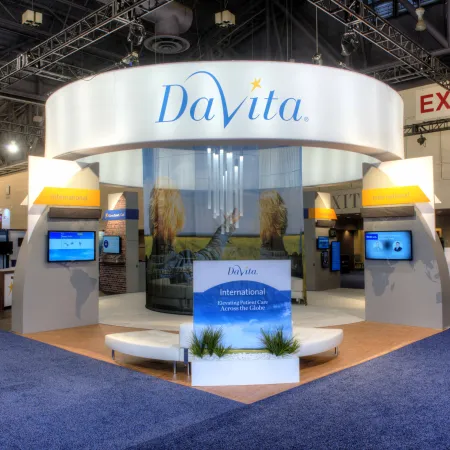 DaVita exhibition booth at a trade show with branded signage, digital screens, and seating area on blue carpet.