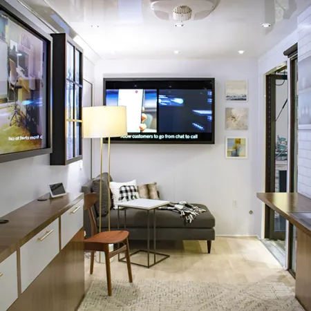 Modern office lounge with gray sofa, wooden chair, floor lamp, large screens, and contemporary decor.