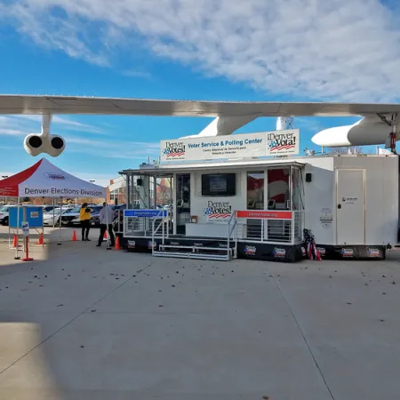 Mobile Denver voter service and polling center set up outdoors under airplane wing with tents and flags nearby.