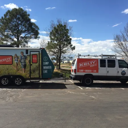 White van towing trailer with Kelty outdoor brand logos parked near trees under a blue sky with clouds.