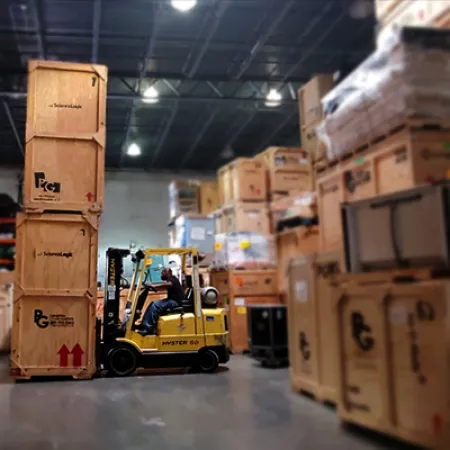 Worker operating yellow forklift moving tall wooden crates inside a warehouse with stacked boxes.