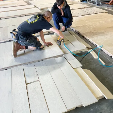 Two men assembling wood panels on a workshop floor with power tools and a forklift in the background
