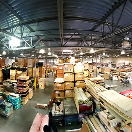 Wide view of a large warehouse filled with stacked boxes, rolls of materials, and industrial shelving under metal roof.
