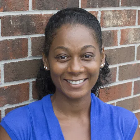 Smiling woman in a blue top with lace detail standing against a red brick wall background.