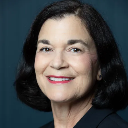 Smiling middle-aged woman with dark hair wearing a black blazer and pearl necklace against a dark background