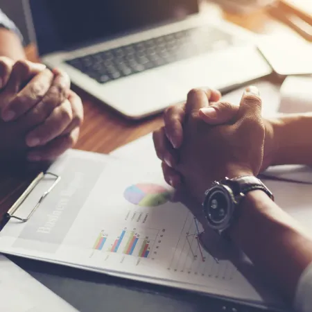 Two people in a business meeting with charts, laptop, and coffee on a wooden table, hands clasped in discussion.