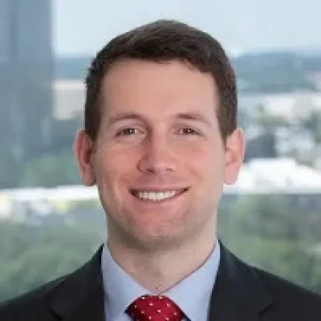 Smiling man in business suit with red polka dot tie against an office window background.