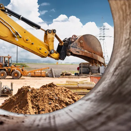 Yellow excavator arm digging soil at a construction site viewed through a large pipe against a cloudy sky.
