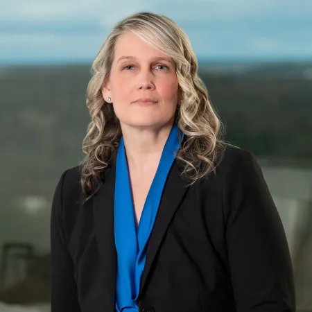 Professional woman with blonde curly hair wearing a black blazer and blue blouse in office setting.