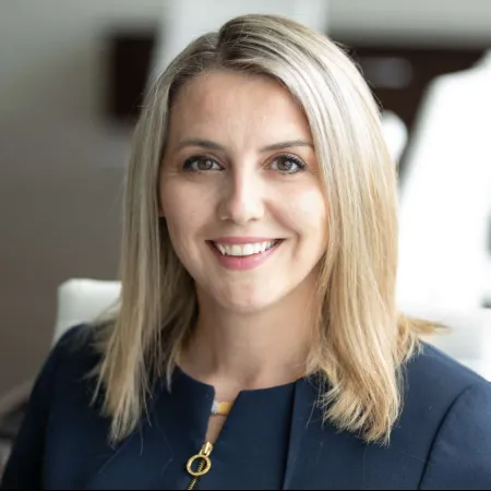 Smiling woman with blonde hair wearing a dark blazer in a bright office setting.