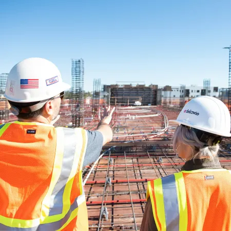 Two construction workers in safety vests and helmets inspecting a building site with rebar and concrete forms.