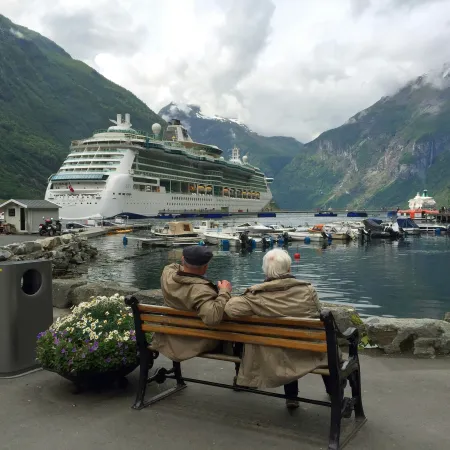 Elderly couple sitting on a bench by a fjord with a cruise ship and boats surrounded by mountains under cloudy sky.