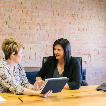Two women in a meeting discussing work with a tablet and notebook, seated at a wooden table with a brick wall background
